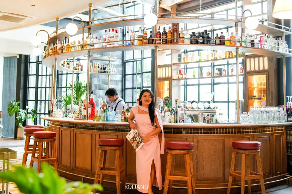 ผู้หญิงในชุดเดรสสีชมพูยืนอยู่หน้าร้านซูชิ Khaolak Sushi ซึ่งมีบาร์รูปวงกลมที่เต็มไปด้วยขวดเครื่องดื่ม ขณะที่บาร์เทนเดอร์กำลังทำงานอยู่หลังเคาน์เตอร์.