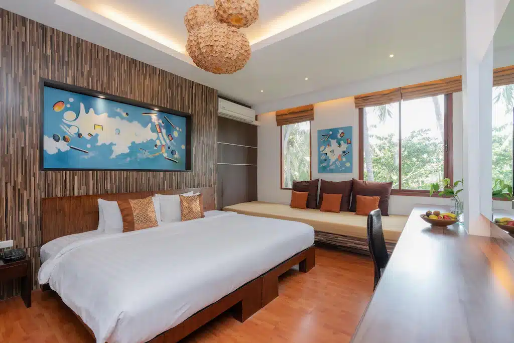 ห้องนอนสไตล์โมเดิร์นพร้อมเตียงขนาดใหญ่, ศิลปะผนังสไตล์สวย, ม้านั่งบิวท์อินพร้อมเบาะ, พื้นไม้, โต๊ะทำงาน, และหน้าต่างขนาดใหญ่ที่ช่วยให้แสงสว่างจากธรรมชาติส่องเข้ามา—เหมาะสำหรับผู้ที่ต้องการที่พักประจวบคีรีขันธ์ที่สะดวกสบายและมีสไตล์.