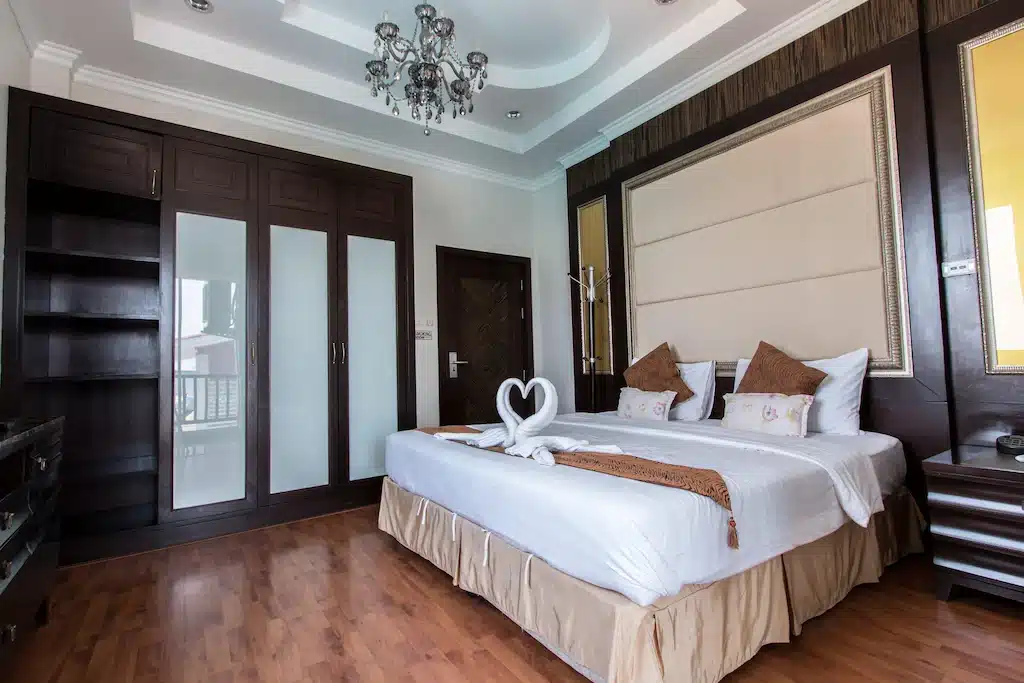 ห้องพักโรงแรมสมัยใหม่พร้อมเตียงขนาดใหญ่ ผ้าขนหนูรูปหงส์ พื้นไม้ ตู้เสื้อผ้าบิวท์อิน โคมไฟระย้า และโต๊ะข้างเตียง—เหมาะสำหรับผู้ที่มองหาที่พักเกาะล้านราคาถูกพร้อมความสะดวกสบายและความหรูหรา.