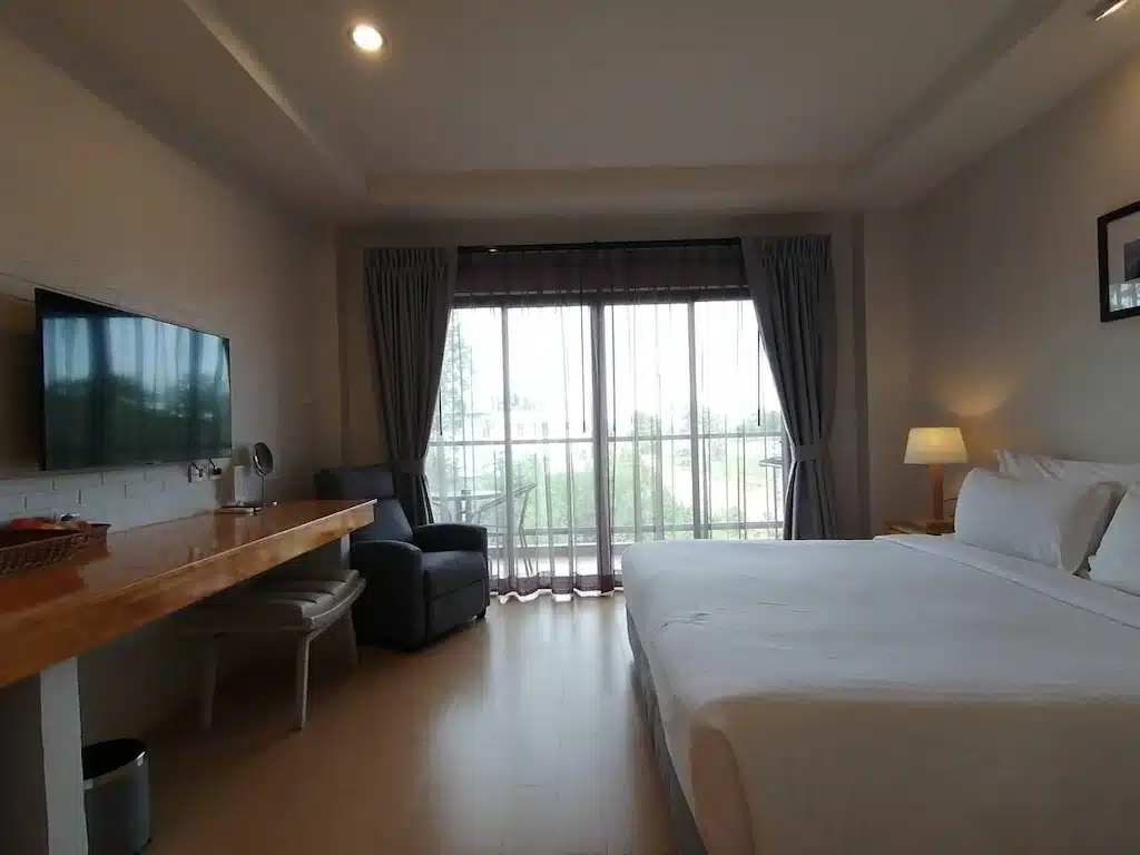ห้องพักโรงแรมปราณบุรีสไตล์โมเดิร์น พร้อมเตียงขนาดใหญ่ โต๊ะทำงานพร้อมเก้าอี้ และโทรทัศน์ เก้าอี้นวมริมหน้าต่าง และผ้าม่านยาวถึงพื้นเพื่อให้แสงสว่างจากธรรมชาติส่องเข้ามา.