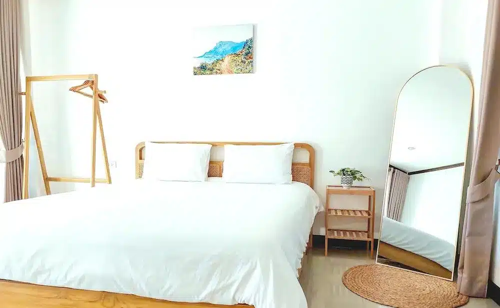 ห้องนอนสไตล์มินิมอลที่ได้แรงบันดาลใจจาก ที่พักเกาะล้านติดทะเล ประกอบด้วยเตียงไม้ ผ้าปูที่นอนสีขาว โต๊ะข้างเตียงพร้อมต้นไม้ กระจกตั้งพื้น ราวแขวนเสื้อผ้า และภาพวาดทิวทัศน์ในกรอบ เพื่อสร้างบรรยากาศเงียบสงบแบบเกาะ.