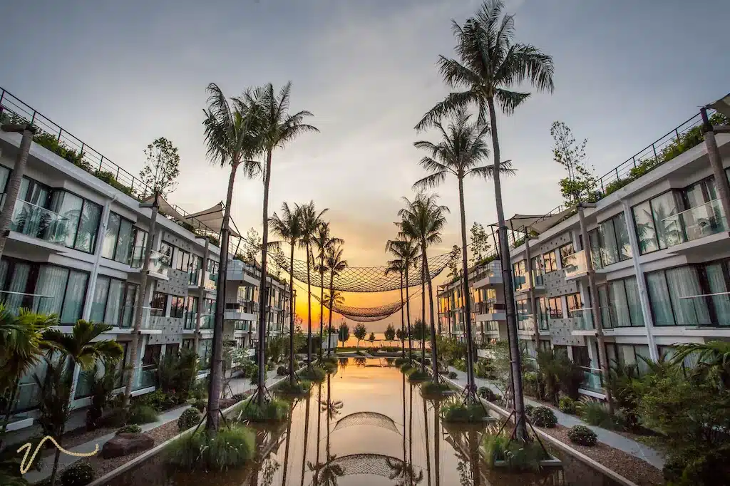 โรงแรมประจวบสมัยใหม่ที่มีต้นปาล์มและสระน้ำสะท้อนแสงอยู่ตรงกลาง ปรากฏในภาพพระอาทิตย์ตกดิน พร้อมทางเดินกลางแจ้งและห้องพักที่มีผนังกระจกทั้งสองด้าน.