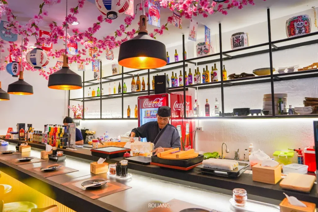 เชฟซูชิเตรียมอาหารอยู่หลังเคาน์เตอร์สมัยใหม่ในร้านอาหารญี่ปุ่นที่สว่างไสว ซึ่งดอกไม้สีชมพู โคมไฟกระดาษ และการตกแต่งที่ได้รับแรงบันดาลใจจากหุบเขาหมอกช่วยสร้างบรรยากาศที่สงบเงียบ.