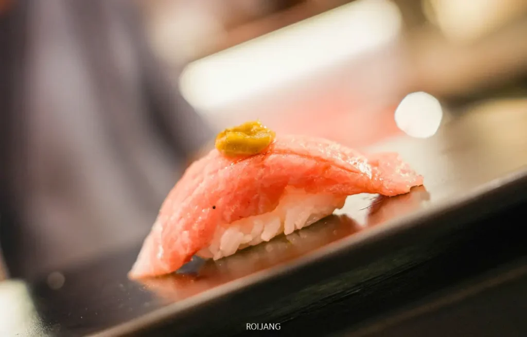 ซูชิชิ้นหนึ่งที่มีเนื้อปลาดิบหั่นบางวางบนวาซาบิเล็กน้อย เสิร์ฟบนจานสีดำที่ชวนให้นึกถึงหุบเขาหมอกอันเงียบสงบ.