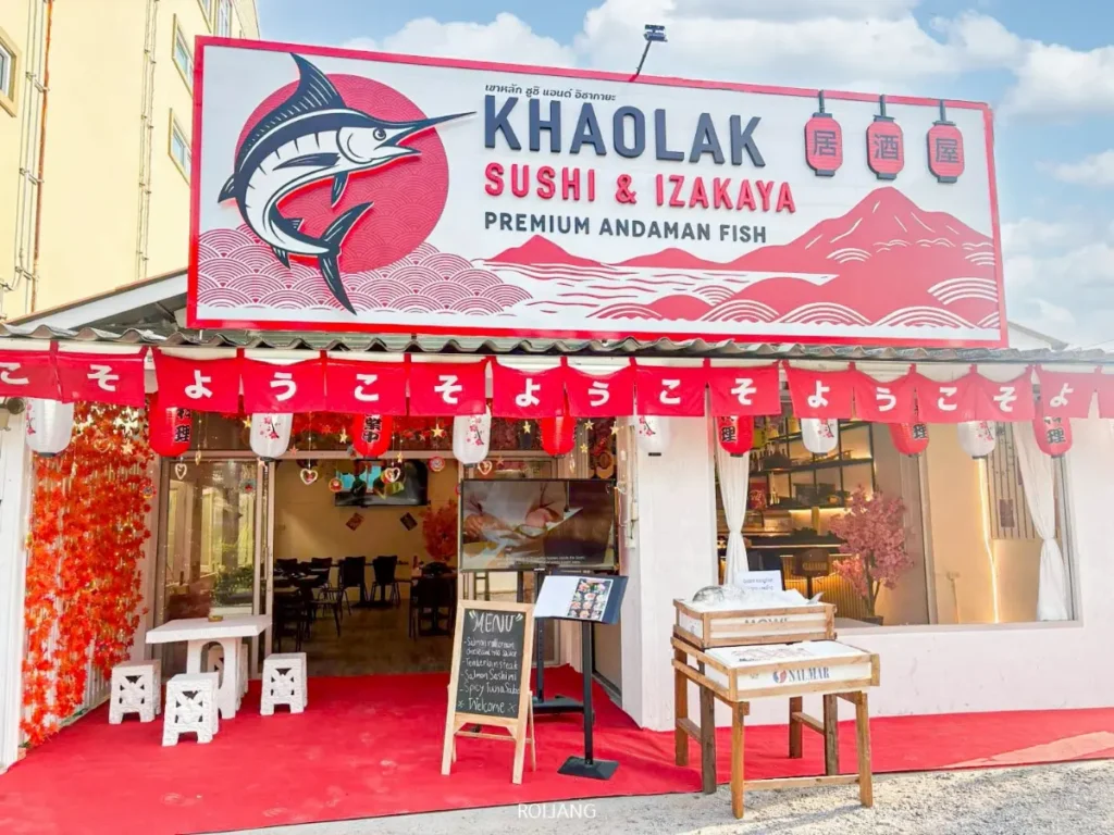 ภายนอกร้านอาหาร Khaolak Sushi & Izakaya ใน Mist Valley โดดเด่นด้วยป้ายสีแดงและขาว โคมไฟญี่ปุ่น ที่นั่งกลางแจ้ง และป้ายเมนูที่ทางเข้า.