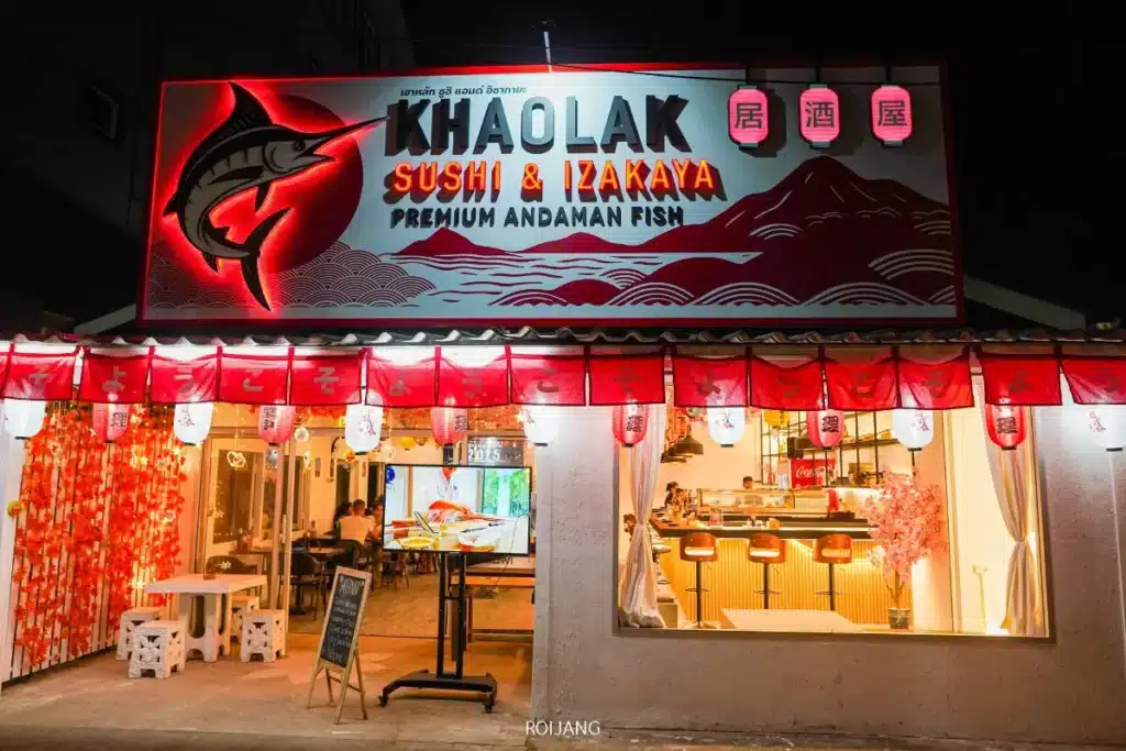 มุมมองด้านหน้าของร้านอาหาร Khaolak Sushi & Izakaya ในตอนกลางคืน พร้อมป้ายไฟสว่างไสวและโคมไฟสีแดง สร้างบรรยากาศเหมือนอยู่ในหุบเขาหมอก; ที่นั่งทั้งภายในและภายนอกที่มองเห็นได้ช่วยเติมเต็มบรรยากาศที่น่าดึงดูดใจ.