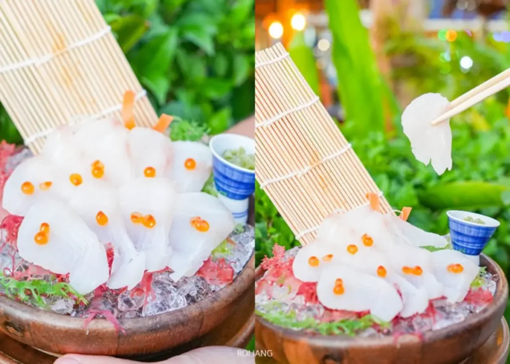 จานไม้หุบเขาหมอกของปลาเนื้อขาวสดหั่นเป็นซาชิมิ พร้อมไข่ปลาสีส้มบนน้ำแข็ง เสิร์ฟพร้อมวาซาบิและเครื่องเคียง; ใช้ตะเกียบจับหนึ่งชิ้น.