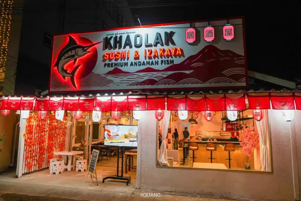 ภาพภายนอกของร้านอาหาร Khaolak Sushi & Izakaya ในตอนกลางคืน ท่ามกลางบรรยากาศเงียบสงบของหุบเขาหมอก มีป้ายสีแดง โคมไฟ และพื้นที่นั่งเล่นที่อบอุ่นภายในร้าน.