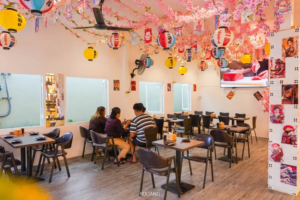 ร้านอาหารในหุบเขาหมอกที่สว่างไสวด้วยการประดับตกแต่งด้วยดอกซากุระและโคมไฟแขวน มีผู้คนหลายคนที่นั่งอยู่ที่โต๊ะ; โต๊ะและเก้าอี้ว่างเปล่าเติมเต็มพื้นที่เหลืออยู่.