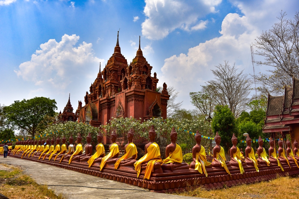 พระพุทธรูปนั่งเรียงรายในชุดจีวรสีเหลืองประดับอยู่ด้านหน้าของวัดสีแดง ท่ามกลางท้องฟ้าที่มีเมฆบางส่วนปกคลุม สะท้อนความงดงามอันเงียบสงบของสถานที่ท่องเที่ยวบุรีรัมย์.