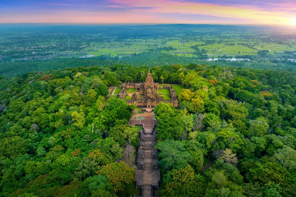 ภาพมุมสูงของกลุ่มวัดโบราณ หนึ่งในสถานที่ท่องเที่ยวบุรีรัมย์ยอดนิยม ล้อมรอบด้วยป่าทึบ มีทางเดินหินยาวทอดไปยังโครงสร้างหลักภายใต้ท้องฟ้าสีสันสดใส.