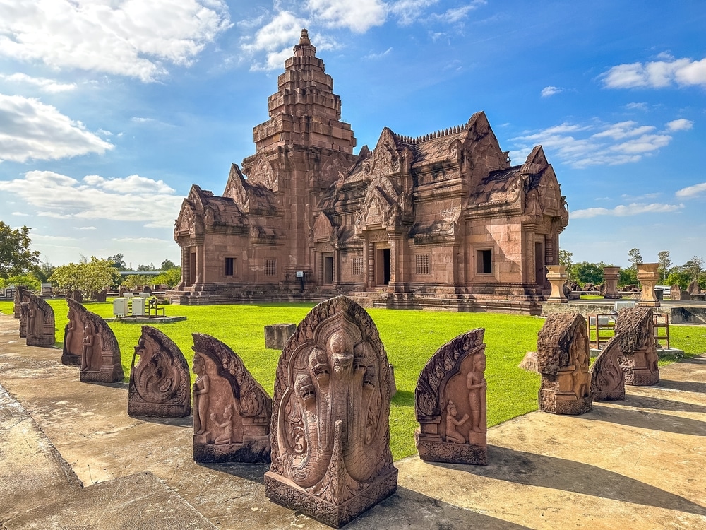 วัดหินโบราณที่มีลวดลายแกะสลักอย่างวิจิตรและแถวของประติมากรรมที่มีรายละเอียดอยู่ด้านหน้า ท่ามกลางท้องฟ้าสีครามที่มีเมฆกระจายอยู่—เป็นหนึ่งในสถานที่ท่องเที่ยวบุรีรัมย์ที่ต้องไปเยือนสำหรับผู้รักวัฒนธรรม.