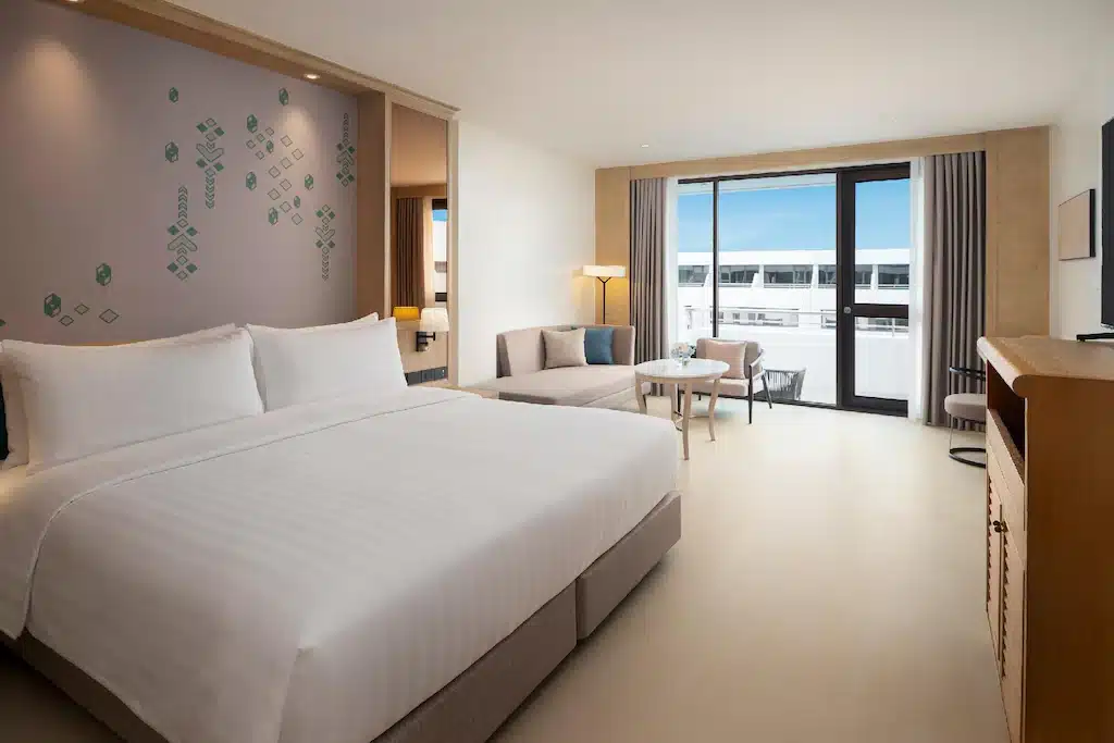 ห้องพักโรงแรมสมัยใหม่พร้อมเตียงขนาดใหญ่ โซฟา โต๊ะกาแฟ และระเบียงพร้อมประตูกระจกที่เปิดรับแสงธรรมชาติ—เหมาะสำหรับการพักผ่อนหลังจากสำรวจสถานที่ท่องเที่ยวในกรุงเทพฯ โทนสีกลางและการตกแต่งแบบมินิมอลสร้างบรรยากาศที่สะอาดและกว้างขวาง.