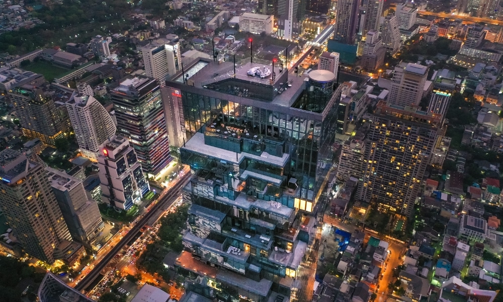 ภาพมุมสูงของทิวทัศน์เมืองสมัยใหม่ในยามพลบค่ำของกรุงเทพฯ ที่โดดเด่นด้วยอาคารกระจกสูงดีไซน์เอกลักษณ์—หนึ่งในสถานที่ท่องเที่ยวกรุงเทพ—รายล้อมไปด้วยตึกระฟ้าและถนนเมืองที่คึกคักมีชีวิตชีวา.