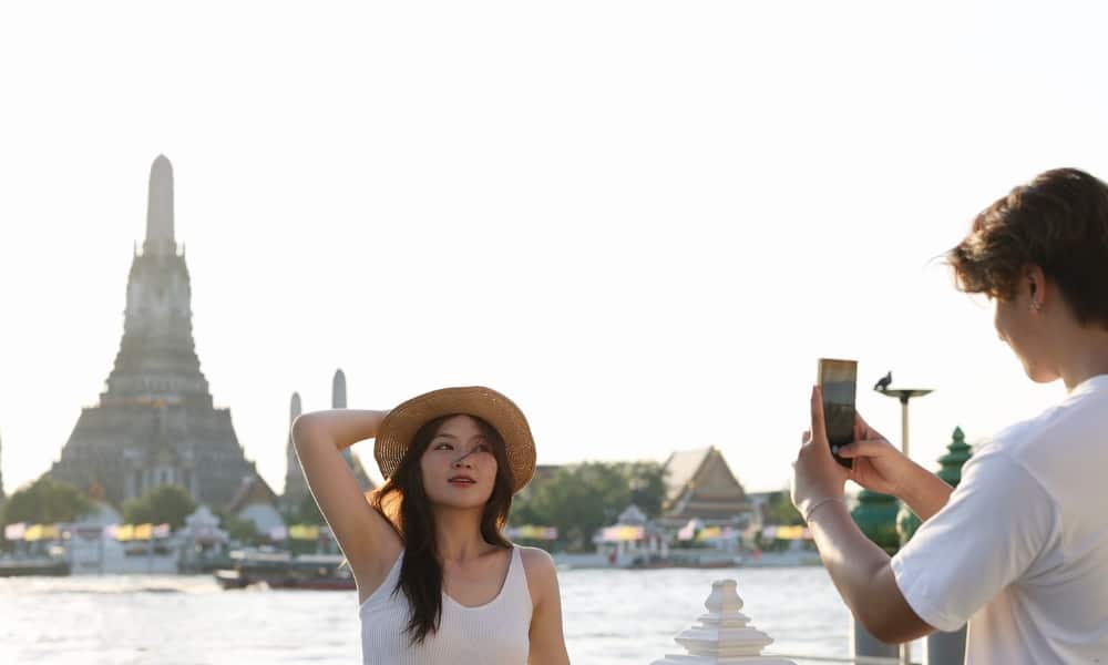 ผู้หญิงคนหนึ่งสวมหมวกฟางโพสท่าถ่ายรูปใกล้แม่น้ำ โดยมีวัดเป็นฉากหลัง—เป็นภาพที่สมบูรณ์แบบในหนึ่งในสถานที่ท่องเที่ยวที่ดีที่สุดในกรุงเทพมหานคร ถ่ายด้วยสมาร์ทโฟน.