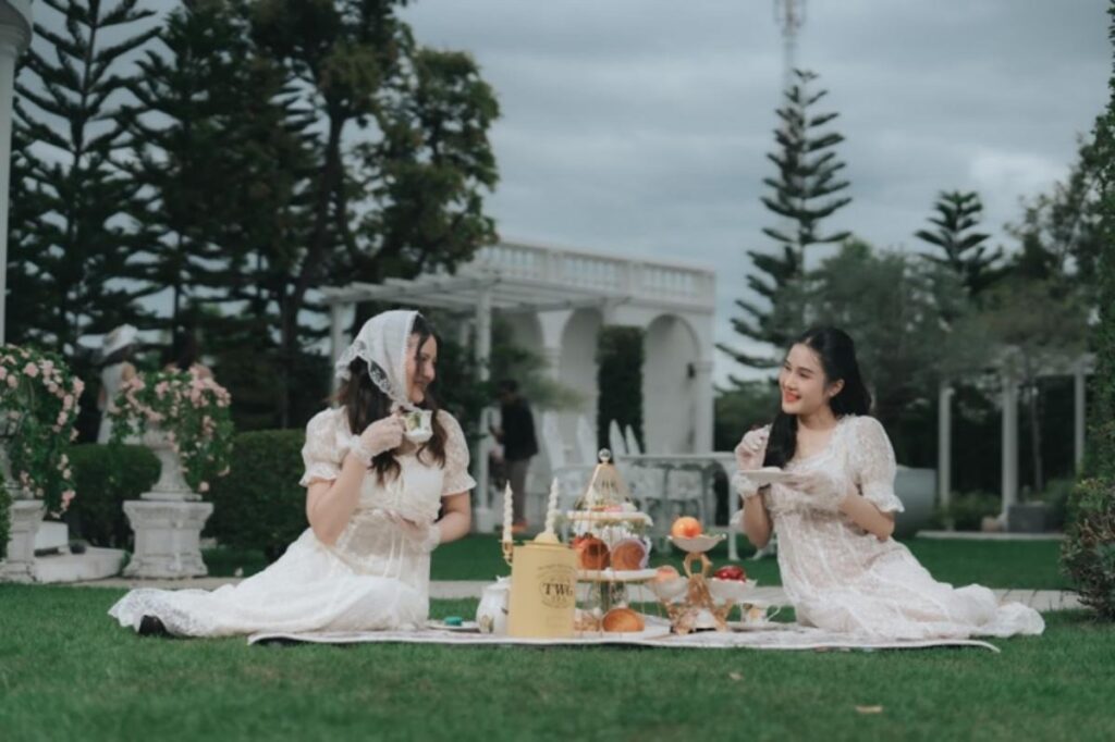 ผู้หญิงสองคนในชุดเดรสสีขาวกำลังดื่มชาและทานขนมปังกับของหวานบนผ้าปูปิกนิกในสวน ล้อมรอบไปด้วยต้นไม้และพืชพรรณสีเขียวภายใต้ท้องฟ้าที่มีเมฆครึ้ม.