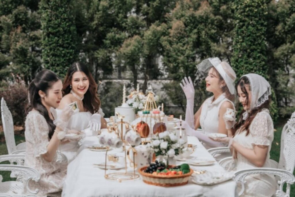 ผู้หญิงสี่คนในชุดเดรสสีขาวสไตล์วินเทจ นั่งอยู่ที่โต๊ะกลางแจ้งซึ่งจัดเตรียมไว้สำหรับดื่มชา ล้อมรอบไปด้วยต้นไม้ใบเขียว และประดับประดาด้วยดอกไม้และของใช้สำหรับชาหลากหลายชนิด.