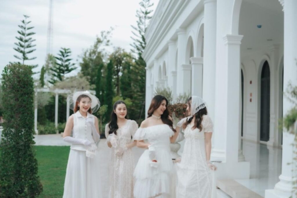 ผู้หญิงสี่คนในชุดสีขาว ยืนและเดินอยู่ภายนอกอาคารสีขาวที่มีซุ้มโค้ง ล้อมรอบด้วยต้นไม้และพืชพรรณสีเขียว.