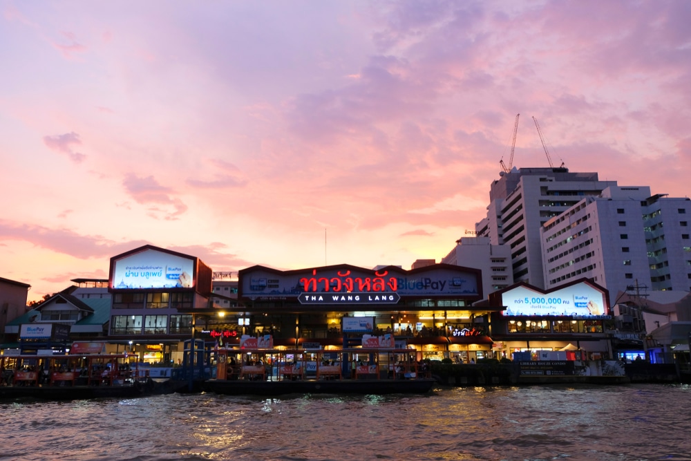 อาคารตลาดริมแม่น้ำที่มีป้ายไฟนีออนสะท้อนแสงยามพระอาทิตย์ตก ทอดเงาบนผืนน้ำตัดกับท้องฟ้าสีชมพูและม่วง—ทัศนียภาพอันน่ามหัศจรรย์ของสถานที่ท่องเที่ยวในกรุงเทพฯ เหมาะอย่างยิ่งสำหรับการเดินเล่นยามเย็น.