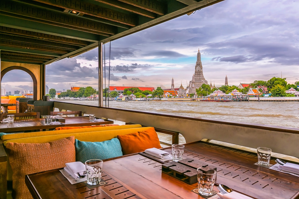 พื้นที่รับประทานอาหารกลางแจ้งของร้านอาหารพร้อมโต๊ะจัดวางอย่างเป็นระเบียบ มองเห็นวิวแม่น้ำและทัศนียภาพอันงดงามของวัดอรุณและเมือง เหมาะอย่างยิ่งสำหรับสถานที่ท่องเที่ยวในกรุงเทพฯ สำหรับผู้ที่ต้องการประสบการณ์การรับประทานอาหารที่น่าจดจำใจกลางกรุงเทพฯ.