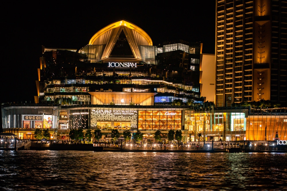 ห้างสรรพสินค้าขนาดใหญ่ที่มีแสงไฟสว่างไสวชื่อว่า ICONSIAM ปรากฏในยามค่ำคืน พร้อมด้วยหน้าร้านแบรนด์หรูและเงาสะท้อนบนผืนน้ำ—เป็นหนึ่งในสถานที่ท่องเที่ยวที่ต้องไปเยือนในกรุงเทพมหานครสำหรับการช้อปปิ้งและชีวิตยามค่ำคืน.