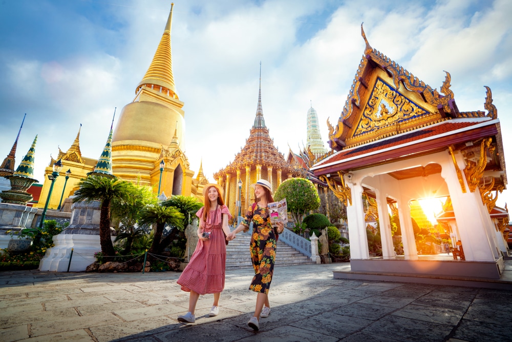 ผู้หญิงสองคนเดินและยิ้มใกล้กับอาคารวัดที่ประดับประดาอย่างสวยงามที่พระบรมมหาราชวัง สถานที่ท่องเที่ยวชั้นนำของกรุงเทพฯ โดยมียอดแหลมสีทองและสถาปัตยกรรมแบบดั้งเดิมเป็นฉากหลัง.