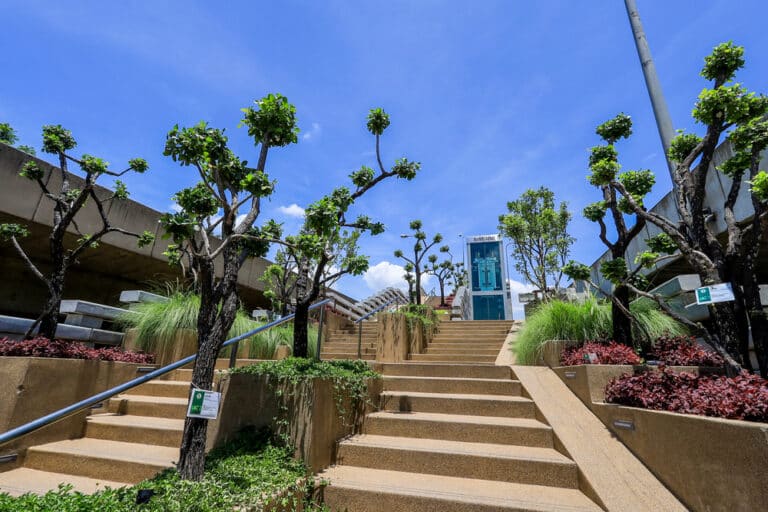 บันไดกลางแจ้งที่เรียงรายไปด้วยต้นไม้ที่ตัดแต่งอย่างสวยงามและพืชพรรณสีเขียว นำทางขึ้นไปยังลิฟต์กระจกใต้ท้องฟ้าสีฟ้าใส ทำให้ที่นี่กลายเป็นจุดพักผ่อนอันเงียบสงบในกรุงเทพฯ สำหรับนักท่องเที่ยวที่ต้องการหลีกหนีความวุ่นวายในเมือง.