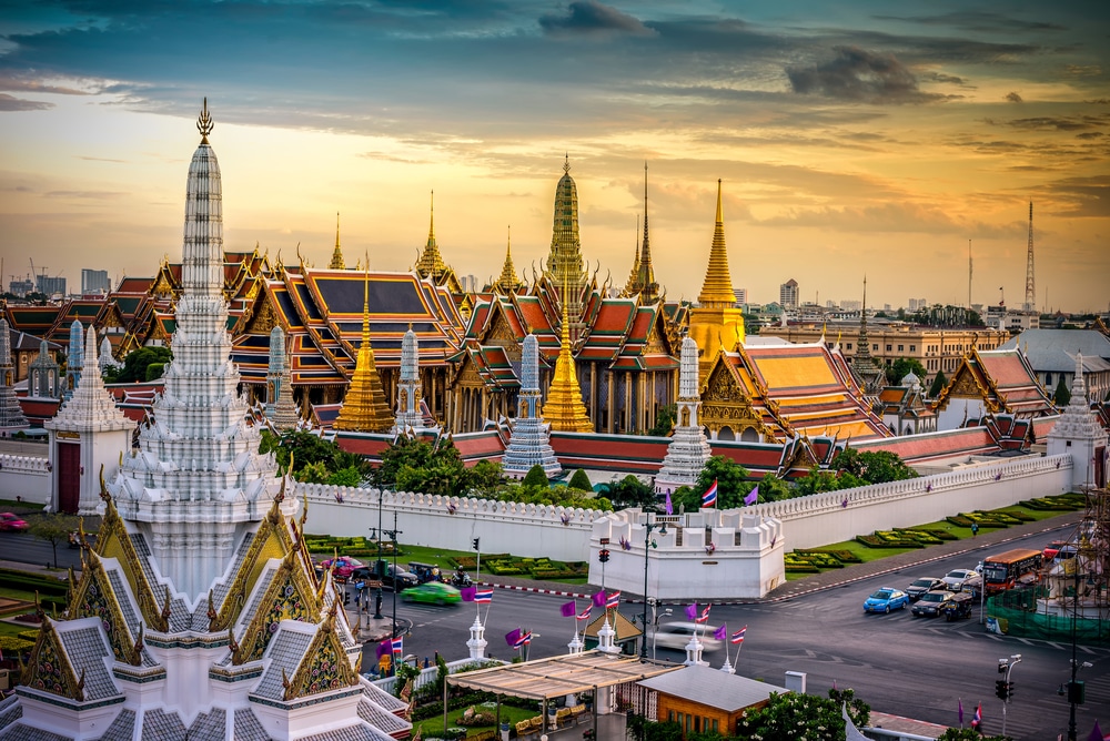 ภาพของพระบรมมหาราชวัง หนึ่งในสถานที่ท่องเที่ยวที่มีชื่อเสียงที่สุดของกรุงเทพฯ โดดเด่นด้วยยอดปราสาททองคำที่ประดับประดาอย่างวิจิตร หลังคาที่ตกแต่งอย่างประณีต และกำแพงสีขาวล้อมรอบในยามพระอาทิตย์ตกดิน.