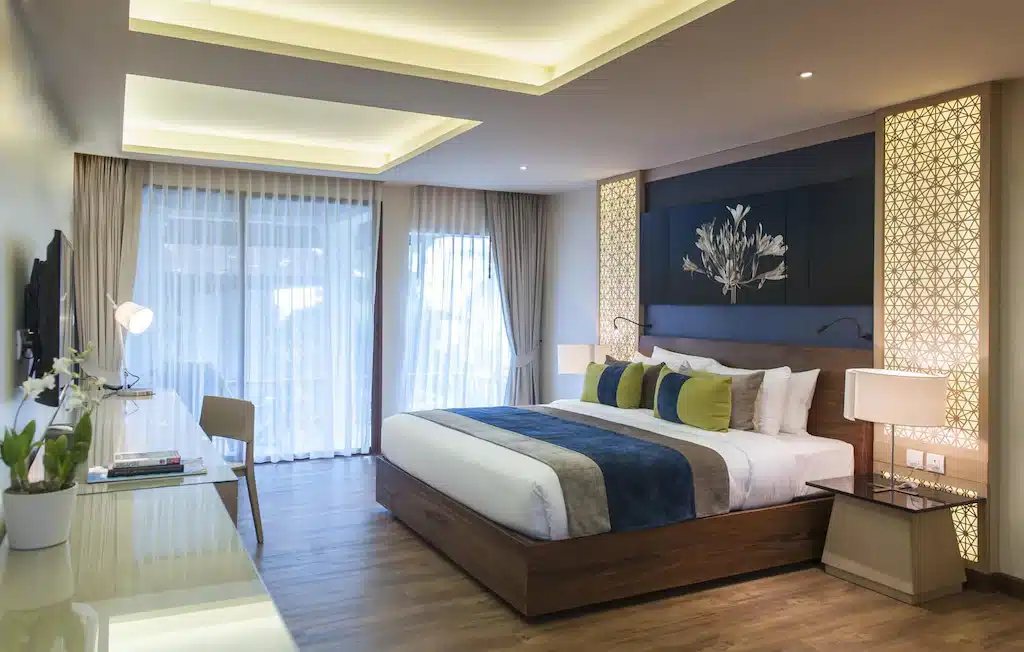ห้องพักโรงแรมสไตล์โมเดิร์น พร้อมเตียงขนาดใหญ่ หมอนตกแต่ง หัวเตียงตกแต่ง โต๊ะทำงาน โทรทัศน์ โคมไฟ และหน้าต่างสูงจากพื้นถึงเพดานพร้อมม่านบาง — เหมาะสำหรับผู้ที่มองหาประสบการณ์ที่พักเกาะสมุยหรือโรงแรมเกาะสมุยที่มีสไตล์.
