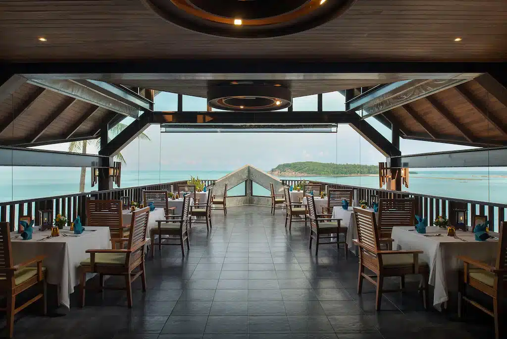 ร้านอาหารกลางแจ้งที่มีเก้าอี้ไม้และผ้าปูโต๊ะสีขาว ให้คุณได้ชมวิวทะเลและเกาะที่อยู่ไกลๆ ผ่านหน้าต่างขนาดใหญ่ใต้เพดานโค้งสูง — เป็นสถานที่รับประทานอาหารที่สมบูรณ์แบบสำหรับแขกที่กำลังมองหา ที่พักสมุย หรือ โรงแรมเกาะสมุย.