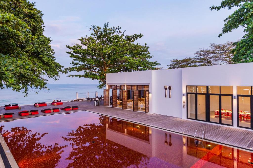 ร้านอาหารริมชายหาดสไตล์ทันสมัยที่มีหน้าต่างขนาดใหญ่ สระว่ายน้ำปูกระเบื้องสีแดง พื้นที่นั่งรับประทานอาหารกลางแจ้ง และวิวทะเล ล้อมรอบด้วยต้นไม้ในช่วงพระอาทิตย์ตกดิน — เป็นสถานที่รับประทานอาหารที่สมบูรณ์แบบ ซึ่งมักพบได้ในโรงแรมเกาะสมุย หรือเป็นส่วนหนึ่งของตัวเลือกที่พักหรูบนเกาะสมุย.