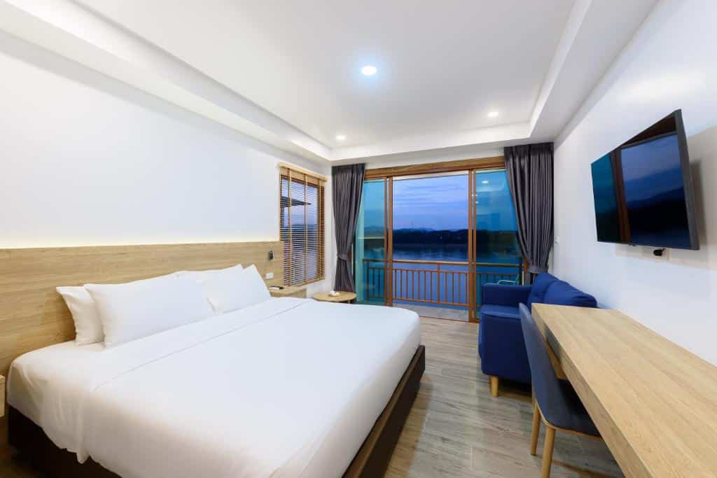 ห้องพักโรงแรมสไตล์โมเดิร์น พร้อมเตียงขนาดใหญ่ เฟอร์นิเจอร์ไม้ โซฟาสีฟ้า ทีวีติดผนัง โต๊ะทำงาน และระเบียงที่มองเห็นวิวผืนน้ำยามพระอาทิตย์ตกดิน — เหมาะอย่างยิ่งสำหรับผู้ที่กำลังมองหา โรงแรมเชียงคาน หรือ ที่พักเชียงคาน ที่ผสมผสานความสะดวกสบายกับวิวทิวทัศน์อันงดงามจนแทบนึกไม่ถึง.