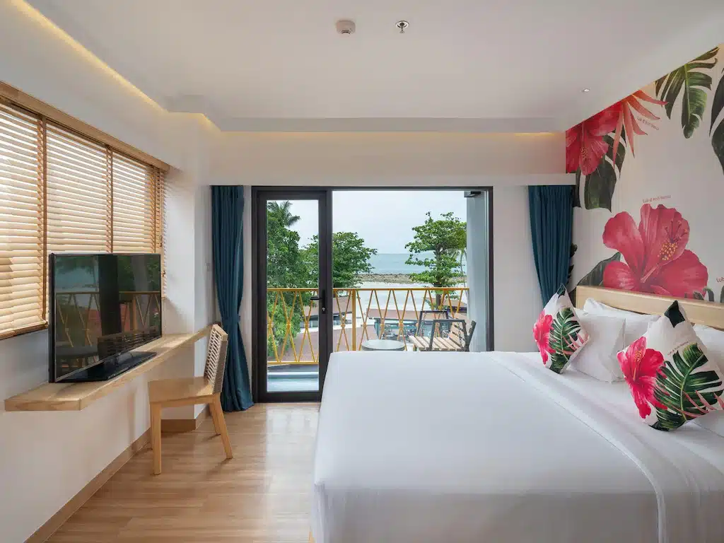 ห้องพักโรงแรมสไตล์ทันสมัย พร้อมเตียงขนาดใหญ่ การตกแต่งด้วยลวดลายดอกไม้ โต๊ะทำงานพร้อมเก้าอี้ ทีวีติดผนัง และระเบียงที่มองเห็นต้นไม้เขตร้อนและทะเล — เหมาะอย่างยิ่งสำหรับแขกที่กำลังมองหาโรงแรมเกาะสมุยที่มีสไตล์ หรือประสบการณ์การพักที่เกาะสมุยที่น่าจดจำ.