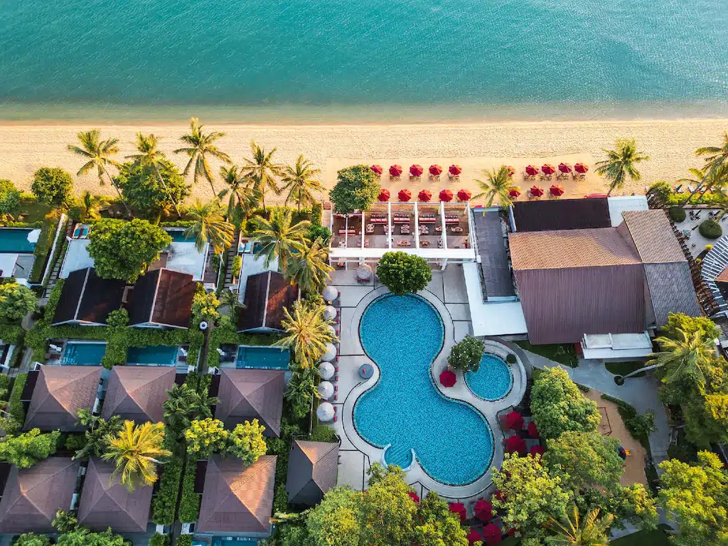 ภาพจากมุมสูงของรีสอร์ทริมชายหาดบนเกาะสมุย ซึ่งมีสระว่ายน้ำกลางรีสอร์ท วิลลาที่ล้อมรอบ ต้นปาล์มที่สั่นไหว และชายหาดทรายขาวที่เรียงรายด้วยร่มสีแดงริมน้ำสีฟ้าใส—เป็นที่พักบนเกาะสมุยที่สมบูรณ์แบบสำหรับผู้ที่ต้องการพักผ่อนในสวรรค์.