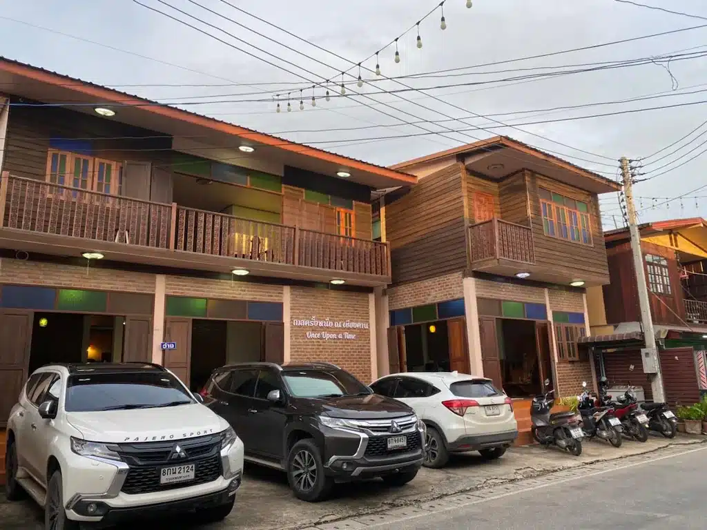 ในช่วงต้นเย็น มีรถ SUV สามคันและรถมอเตอร์ไซค์หลายคันจอดอยู่หน้าอาคารไม้สองชั้นที่มีระเบียงและไฟประดับแขวนอยู่ด้านบน สร้างบรรยากาศที่อบอุ่นและเป็นมิตร ซึ่งมักพบได้ที่พักในเชียงคาน สำหรับนักท่องเที่ยวที่มาสำรวจเมืองริมแม่น้ำที่เต็มไปด้วยเสน่ห์แห่งนี้.