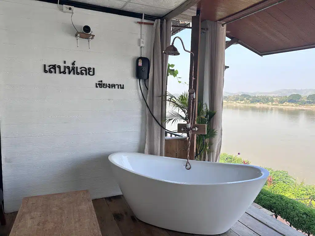 อ่างอาบน้ำแบบตั้งอิสระในห้องน้ำกลางแจ้งที่มีวิวแม่น้ำ ฝักบัวติดผนัง และข้อความภาษาไทยบนผนังสีขาว — เป็นจุดเด่นที่ดึงดูดใจที่โรงแรมเชียงคาน สำหรับผู้ที่กำลังมองหาที่พักเชียงคานราคาถูก ท่ามกลางธรรมชาติที่สวยงามตระการตา.