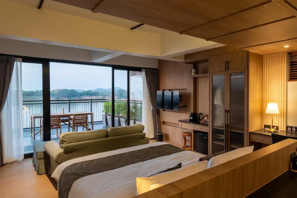 ห้องพักโรงแรมสไตล์ทันสมัย พร้อมเตียงขนาดใหญ่ โซฟา โทรทัศน์ ตู้เย็นขนาดเล็ก และโคมไฟโต๊ะทำงาน มีหน้าต่างสูงจากพื้นถึงเพดานและระเบียงพร้อมที่นั่งกลางแจ้ง มองเห็นวิวทะเลสาบและภูเขาที่สวยงาม—เหมาะสำหรับนักท่องเที่ยวที่ต้องการสำรวจ ที่เที่ยวเชียงคาน หรือค้นพบจุดท่องเที่ยวที่ดีที่สุดในบริเวณใกล้เคียง.