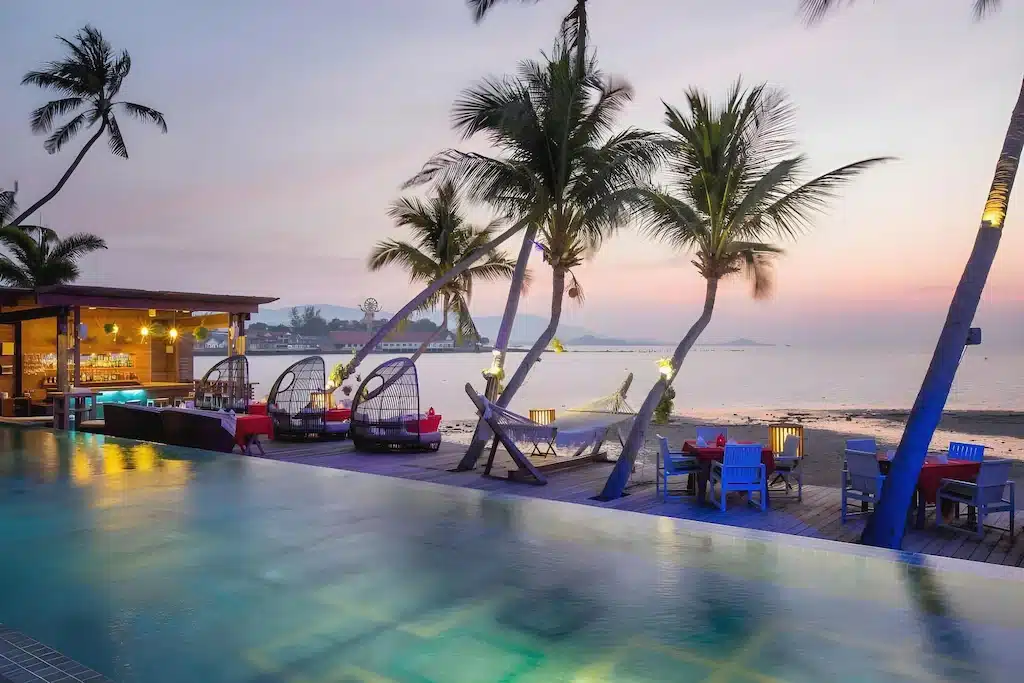 รีสอร์ทริมชายหาดที่มีสระว่ายน้ำ ต้นปาล์ม เตียงห้อย เก้าอี้นอน และโต๊ะรับประทานอาหารในยามพระอาทิตย์ตกดิน — เหมาะอย่างยิ่งสำหรับการพักผ่อนอย่างสบายใจที่ ที่พักเกาะสมุย.