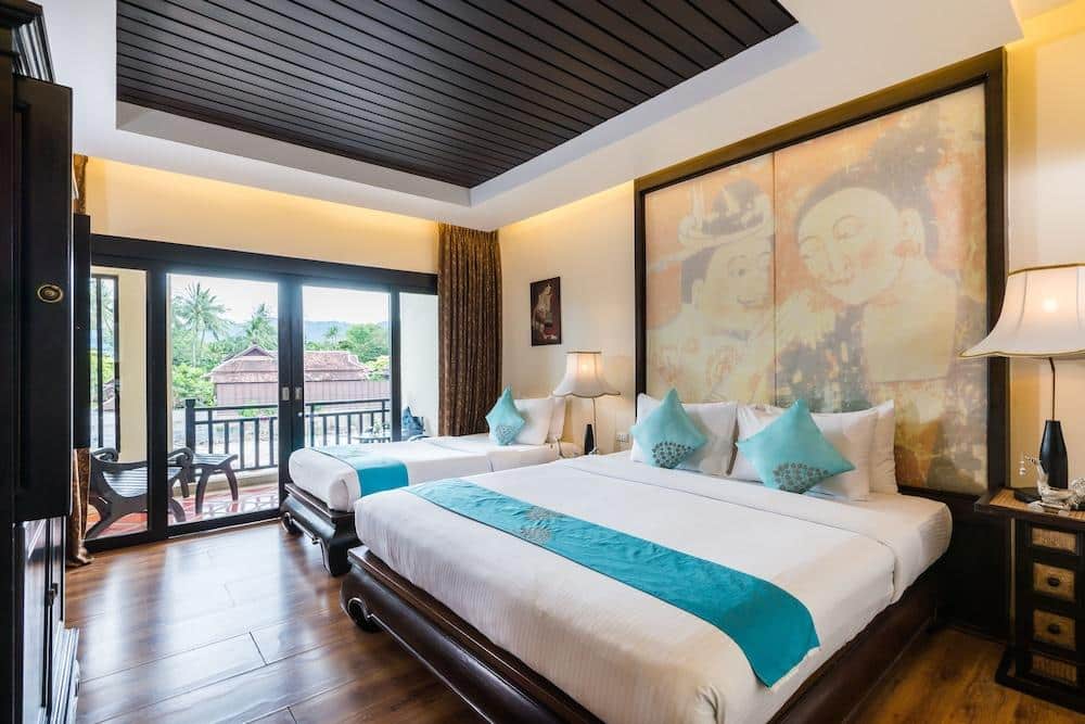 ห้องพักในโรงแรมที่มีเตียงสองเตียง ตกแต่งด้วยสีเทอร์ควอยซ์ มีภาพศิลปะขนาดใหญ่บนผนัง โคมไฟตั้งพื้น และระเบียงที่มีที่นั่งกลางแจ้งพร้อมวิวทิวทัศน์ของพื้นที่สีเขียว — เหมาะอย่างยิ่งสำหรับนักท่องเที่ยวที่มองหาประสบการณ์ที่พักเกาะสมุยหรือโรงแรมเกาะสมุยที่สงบเงียบ ท่ามกลางความงามของธรรมชาติ.