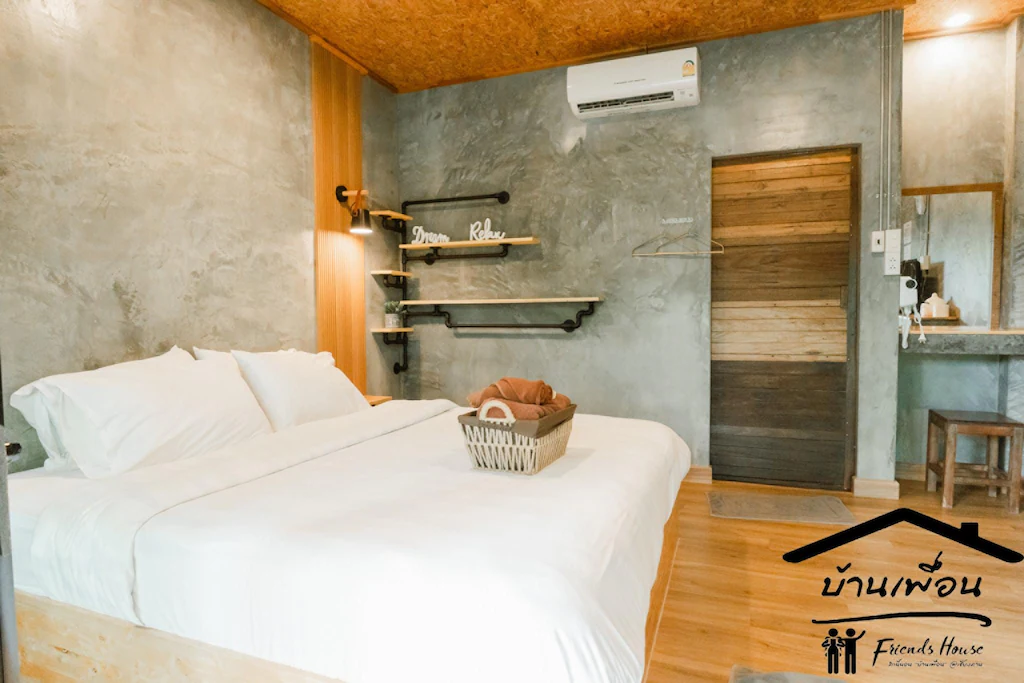 ห้องนอนสไตล์โมเดิร์นที่มีเตียงคู่ ชุดเครื่องนอนสีขาว ผนังคอนกรีต รายละเอียดตกแต่งด้วยไม้ ชั้นวางของ และตะกร้าบนเตียง เหมาะสำหรับผู้ที่กำลังมองหาที่พักเชียงคานราคาถูกที่มีสไตล์ทันสมัย มีโลโก้ Friends House อยู่มุมขวาล่าง.