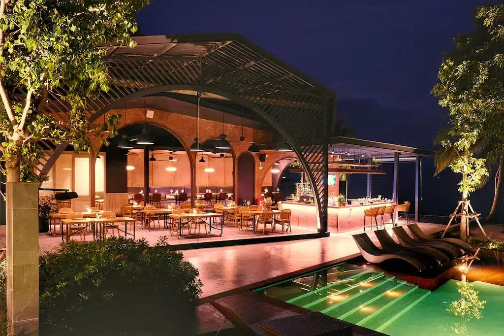ร้านอาหารกลางแจ้งที่มีสถาปัตยกรรมสมัยใหม่ แสงไฟที่อบอุ่น เก้าอี้เลานจ์ และสระว่ายน้ำ ล้อมรอบด้วยต้นไม้ในยามค่ำคืน — เหมาะอย่างยิ่งสำหรับแขกที่ต้องการประสบการณ์การรับประทานอาหารที่ลืมไม่ลง ระหว่างการเข้าพักที่ โรงแรมเกาะสมุย.