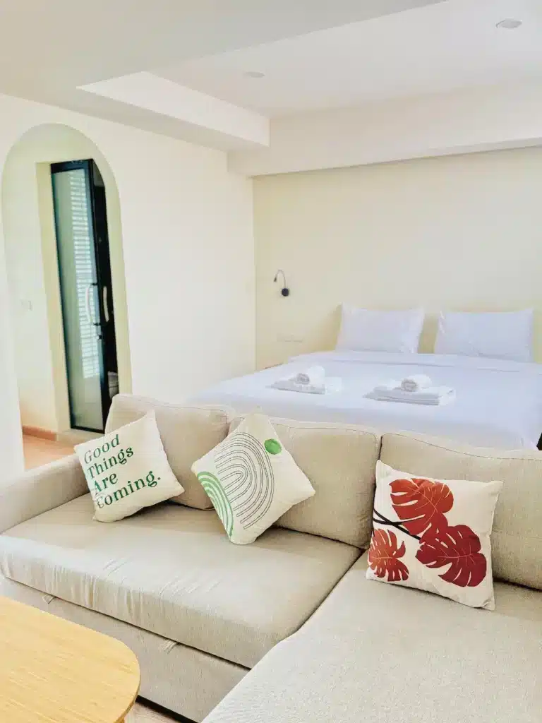 ห้องนอนสไตล์โมเดิร์นและมินิมัลที่ โรงแรมเชียงคาน มีโซฟาสีเบจ หมอนตกแต่งสามใบ โต๊ะไม้ และเตียงที่จัดไว้อย่างเรียบร้อยด้วยผ้าปูเตียงสีขาวและผ้าขนหนูที่ม้วนไว้.