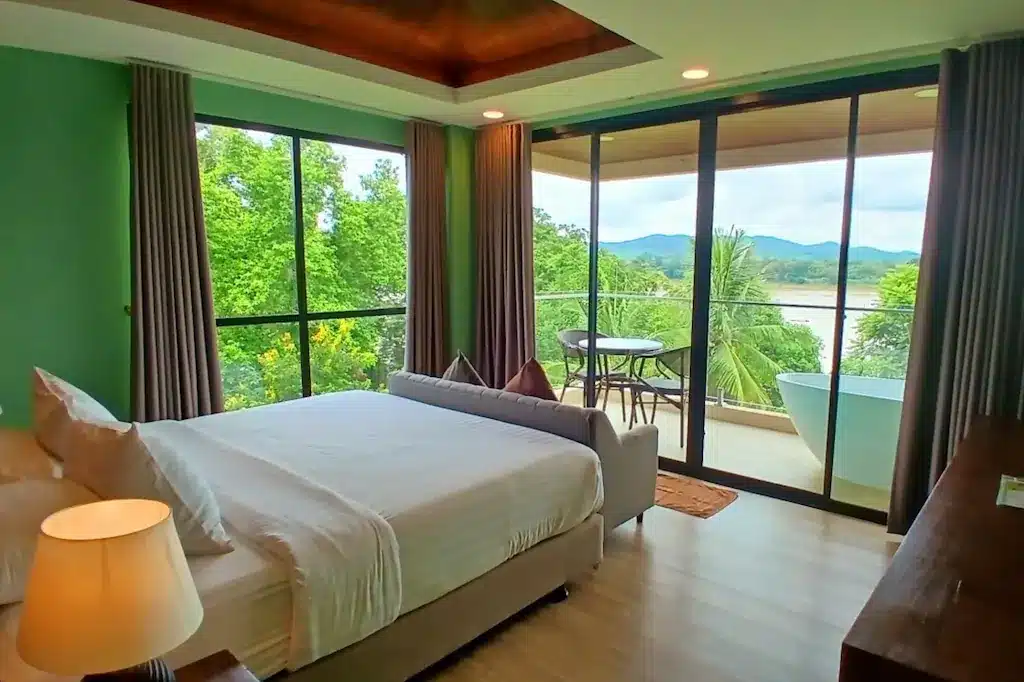 ห้องนอนสไตล์โมเดิร์นที่มีเตียงขนาดใหญ่ โคมไฟ และระเบียงพร้อมที่นั่งกลางแจ้งและอ่างอาบน้ำ มองเห็นต้นไม้เขียวขจีและภูเขาไกลๆ ผ่านหน้าต่างสูงจากพื้นถึงเพดาน — เป็นที่พักที่สมบูรณ์แบบสำหรับผู้ที่ต้องการเที่ยวเชียงคาน หรือสำรวจสถานที่ท่องเที่ยวในเชียงคานอย่างหรูหราและสะดวกสบาย.