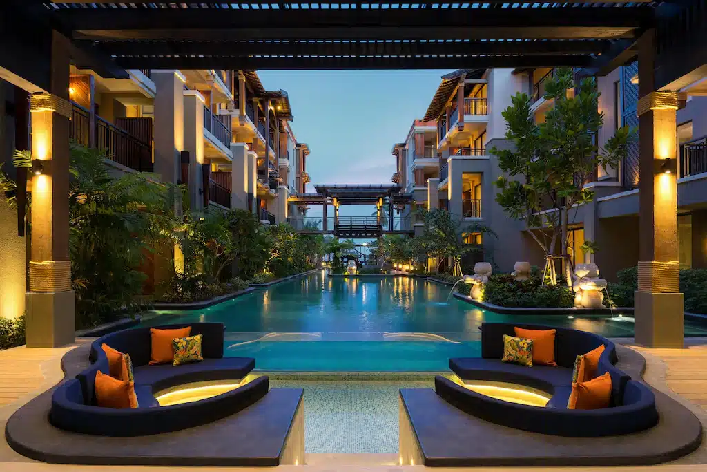 ลานกลางโรงแรมที่ดูสง่างามในยามพลบค่ำ พร้อมพื้นที่นั่งแบบฝังพื้นและหมอนตกแต่งที่หันหน้าสู่สระว่ายน้ำยาว ซึ่งรายล้อมด้วยพืชพรรณและส่องสว่างด้วยแสงไฟสีอบอุ่น สร้างบรรยากาศที่ชวนให้ผ่อนคลายสำหรับแขกผู้แสวงหาประสบการณ์โรงแรมสมุยหรือที่พักเกาะสมุยที่สงบเงียบ.