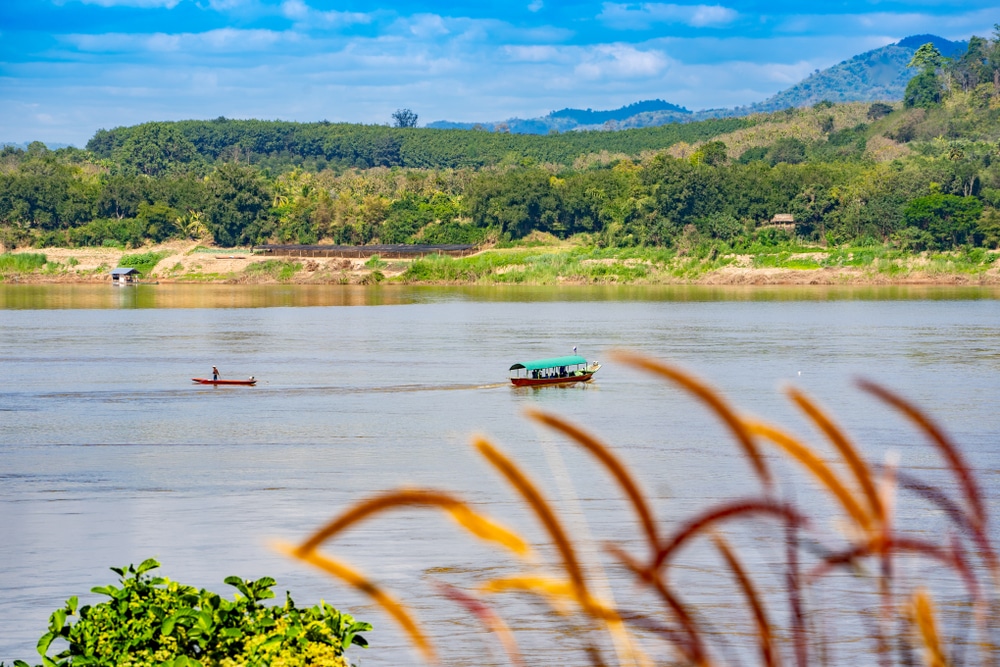 แม่น้ำกว้างที่มีเรือสองลำลอยอยู่บนน้ำ ต้นไม้สีเขียวและภูเขาเป็นฉากหลัง ส่วนด้านหน้ามีหญ้าสูงใต้ท้องฟ้าสีฟ้า—ภาพที่สงบนี้สะท้อนความงามตามธรรมชาติของ เชียงคานที่เที่ยว ซึ่งเหมาะอย่างยิ่งสำหรับผู้ที่ต้องการสัมผัสความงามของการเที่ยวเชียงคาน.