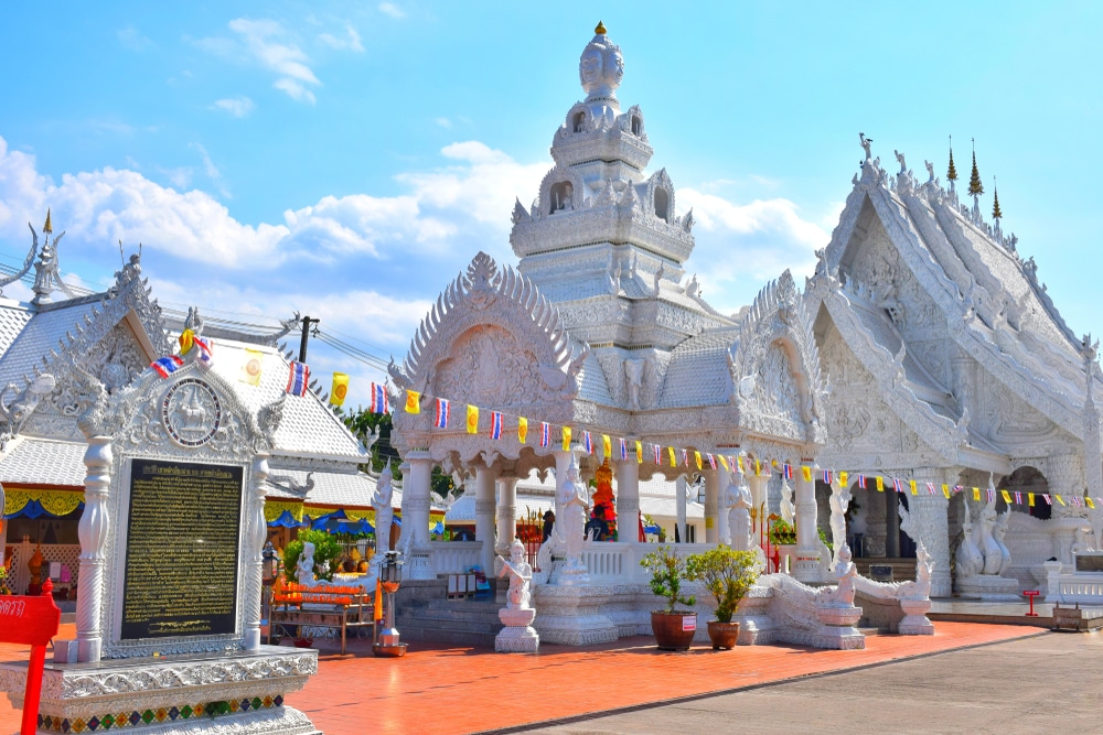 วัดสีขาวที่มีลวดลายแกะสลักและรูปปั้นอันวิจิตรตระการตา ตั้งอยู่ใต้ท้องฟ้าสีคราม ธงหลากสีสันแขวนอยู่เหนือลานที่มีกระเบื้องหลังคาสีแดง ป้ายขนาดใหญ่ตั้งอยู่ด้านหน้า ระบุให้สถานที่ท่องเที่ยวแห่งนี้ในจังหวัดน่าน เป็นหนึ่งในสถานที่ท่องเที่ยวที่น่าประทับใจที่สุดสำหรับนักท่องเที่ยวที่มาเยือนภูมิภาคนี้.