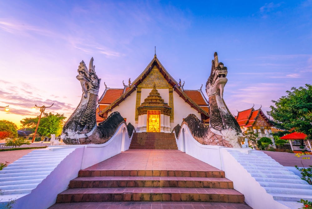 วัดที่ตกแต่งอย่างวิจิตรด้วยหลังคาสีแดงและรูปปั้นมังกรประดับสองข้างบันได ถ่ายภาพในช่วงพระอาทิตย์ตกดิน ท่ามกลางท้องฟ้าสีฟ้าและสีชมพู — เป็นสถานที่ท่องเที่ยวในน่านที่ไม่ควรพลาด สำหรับใครก็ตามที่ต้องการสัมผัสความงามอันเป็นเอกลักษณ์ของการเที่ยวน่าน.