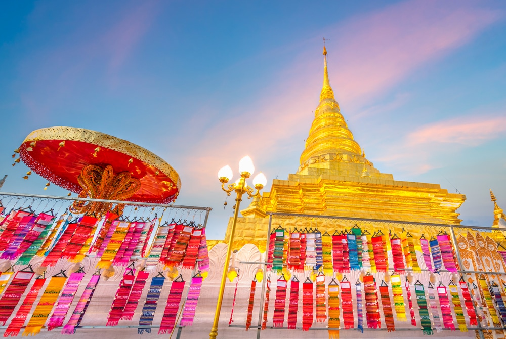 เจดีย์สีทองและวัดที่ประดับด้วยผ้าสีสันสดใส พร้อมร่มสีแดงตั้งอยู่ด้านหน้า ซึ่งถูกแสงไฟส่องสว่างยามพลบค่ำใต้ท้องฟ้าสีฟ้าและสีชมพู—เป็นภาพที่งดงามตระการตา ซึ่งถ่ายทอดเสน่ห์ของ เที่ยวน่าน ได้อย่างสมบูรณ์แบบ ซึ่งเป็นหนึ่งในสถานที่ท่องเที่ยวที่สวยงามที่สุดของ เที่ยวน่าน สำหรับนักท่องเที่ยวที่แสวงหาความมหัศจรรย์ทางวัฒนธรรม.
