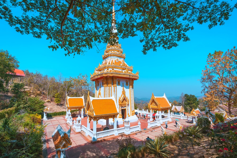 วัดไทยหลังคาทองที่มีรายละเอียดประดับตกแต่งอย่างวิจิตร ตั้งอยู่ใต้ต้นไม้ ล้อมรอบด้วยระเบียงและนักท่องเที่ยว ท่ามกลางท้องฟ้าสีครามและภูมิทัศน์ภูเขา—เหมาะอย่างยิ่งสำหรับผู้ที่กำลังมองหาที่เที่ยวเชียงคาน และต้องการสัมผัสเสน่ห์ของเชียงคานที่เที่ยว.