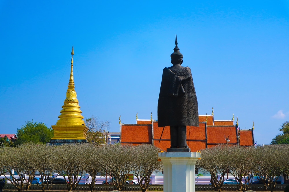 รูปปั้นของบุคคลที่ยืนอยู่บนแท่น พร้อมด้วยเจดีย์สีทอง หลังคาวัด ต้นไม้ และท้องฟ้าสีฟ้าเป็นฉากหลัง — ฉากที่งดงามราวกับภาพวาด ซึ่งถ่ายทอดแก่นแท้ของ “เที่ยวน่าน” และเชิญชวนนักท่องเที่ยวมาค้นพบสถานที่ท่องเที่ยวในน่านเพิ่มเติม.